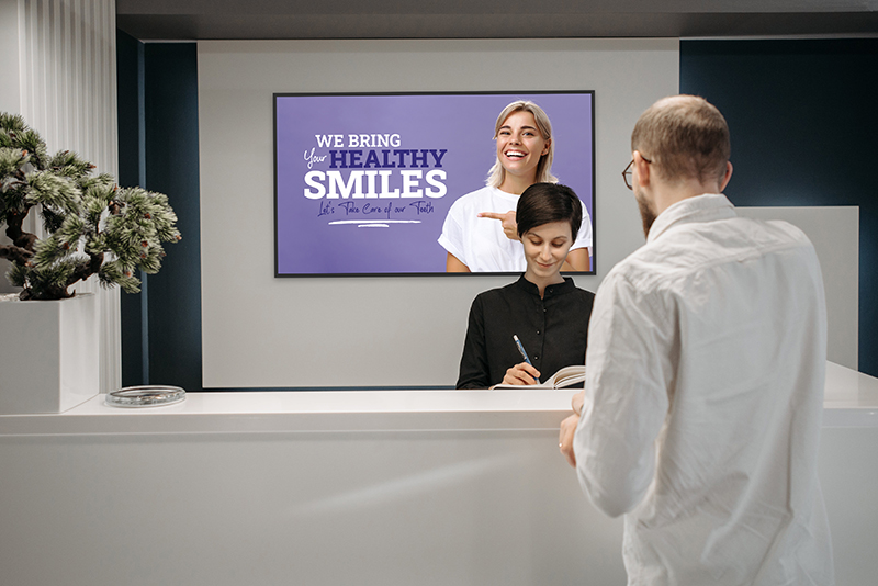 Landscape digital signage screen on the wall behind a reception desk while a customer is served