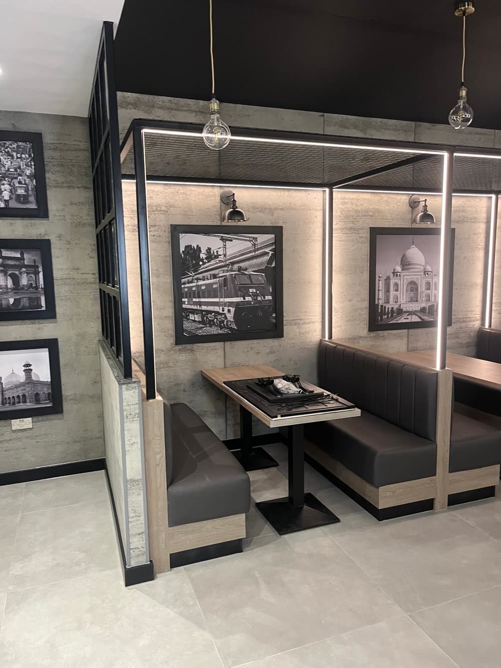Finished restaurant booths: grey upholstered banquettes, oak-effect tables, a black steel frame with LED strip lighting and framed black and white photographs on a concrete-effect wall